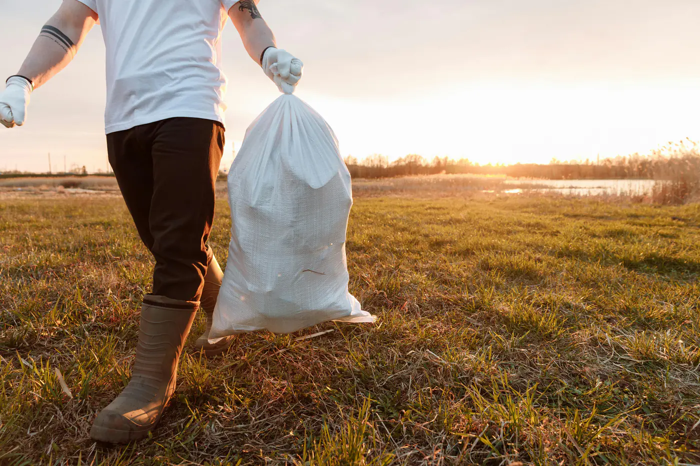 Worker carrying a trash bag through a rural field at sunset for garbage collection