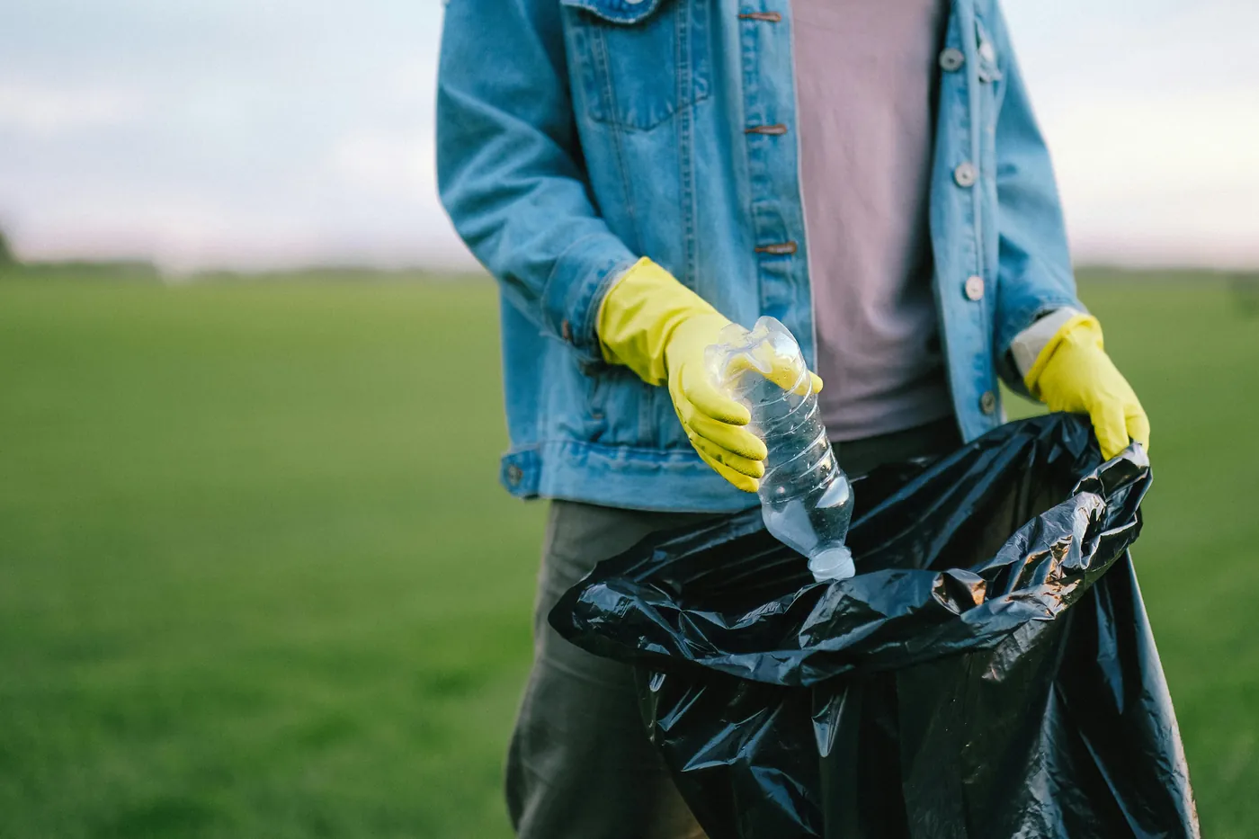 Person in denim jacket and yellow gloves picking up litter in a rural field